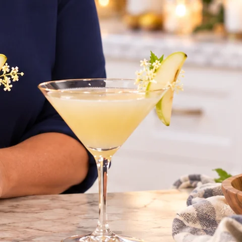 A pale gold pear martini in a stemmed glass, garnished with a slice of pear and elderflower.